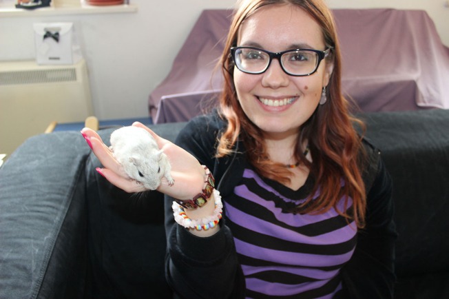 A smiling girl with long red hair and Gok Wan glasses. She is wearing a black and purple striped t-shirt under an open black hoodie. She is holding a grey gerbil in her right hand and holding it up.